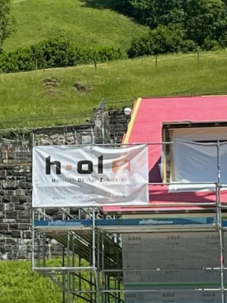 neben Kirche in Schoppernau eingerüstetes Haus mit Banner von Heinrich Olivier Zimmerei, Umbau und Sanierung